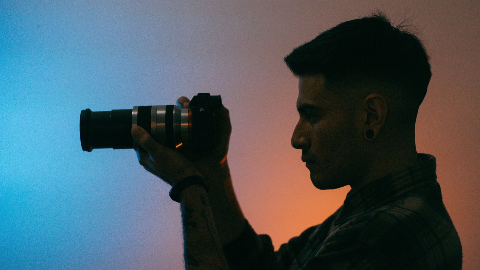 Joshua de perfil, a contraluz, sosteniendo una cámara frente a una pared azul y naranja